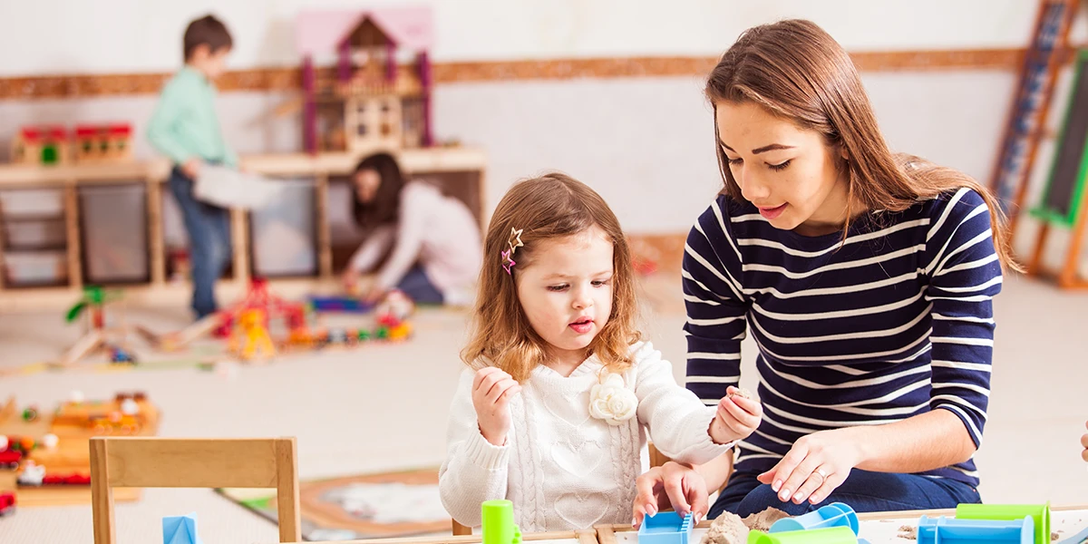 Teacher is teaching the preschooler girl to make sand shapes while other kids are playing together