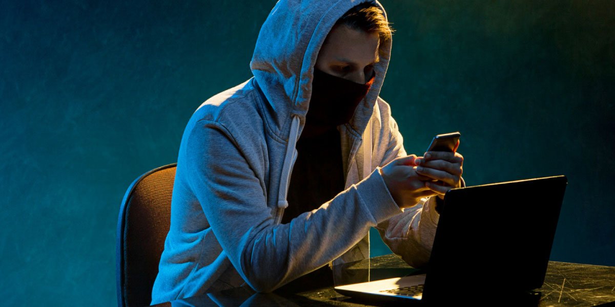 Masked man looking at his phone with his laptop in front of him in a dark room