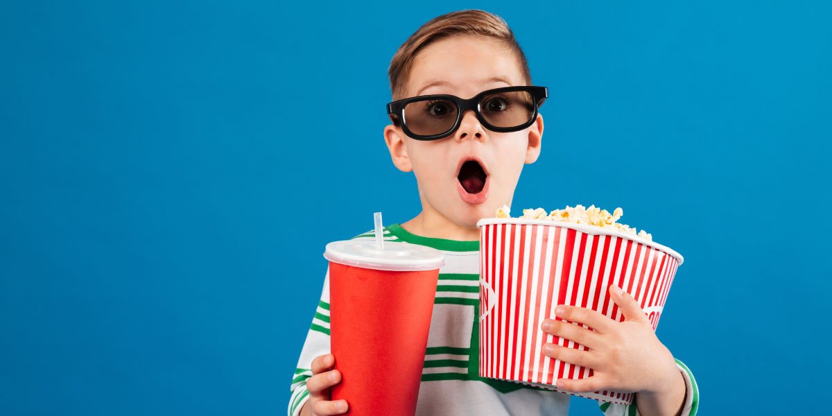 Gaping young boy with sunglasses holding a bucket of popcorn and a cup of coke