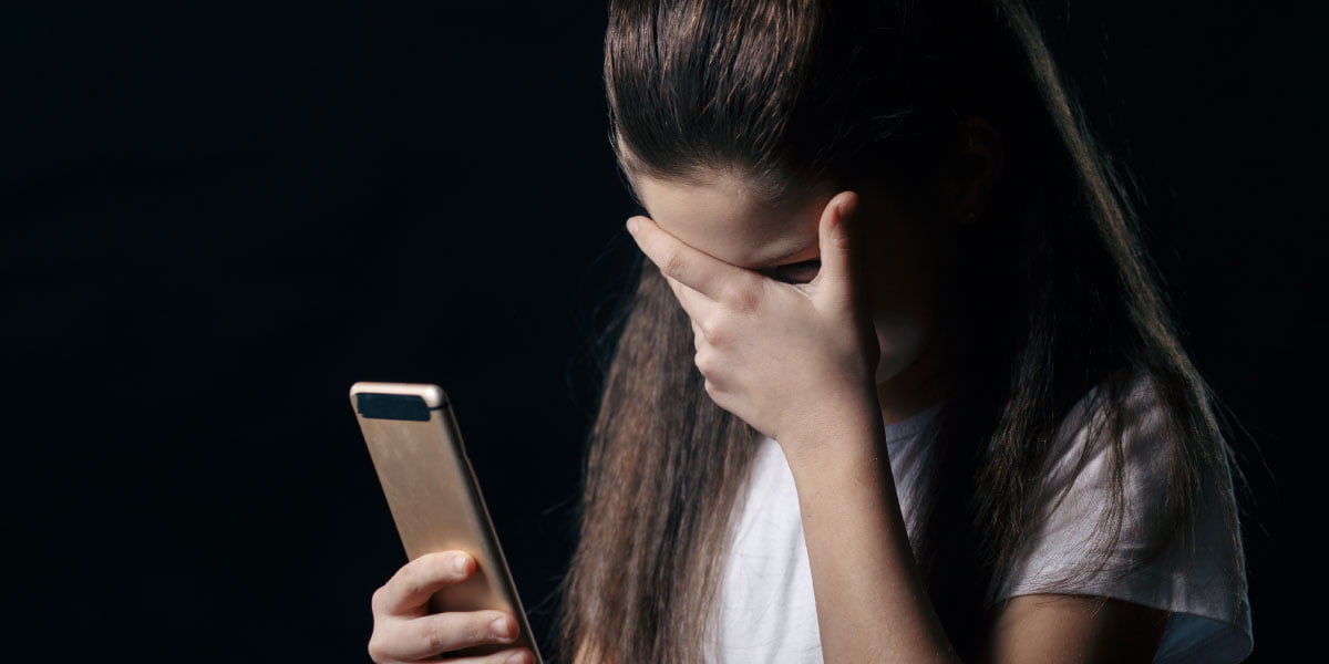 A cyberbullied girl holding a phone with one hand and covering her eyes with the other