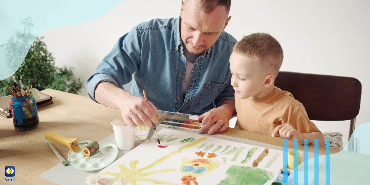 Father using paintbrush and paints to help his little son to paint a picture on paper at the table