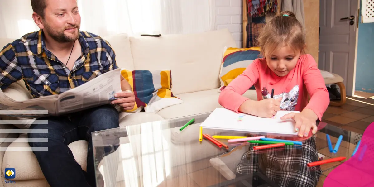 a father sitting on the couch reading a newspaper and spending quality time with her daughter while she’s painting in her notebook