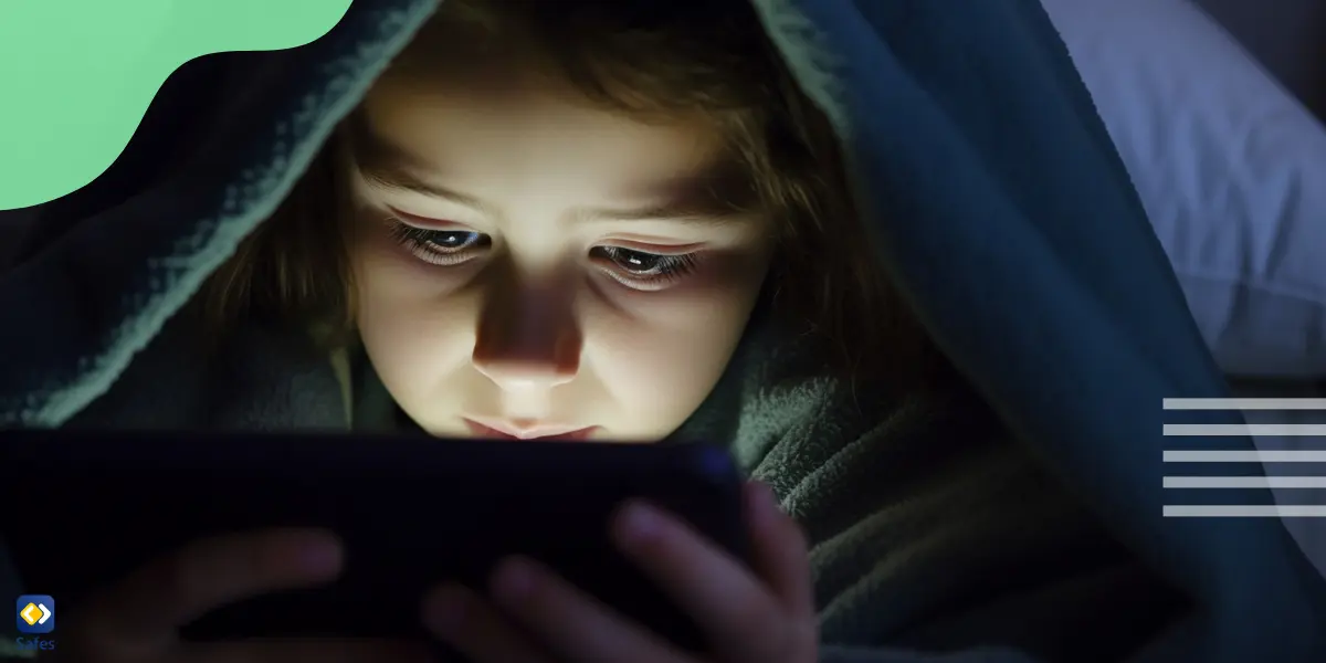 A young girl under the blanket, holding a tablet with both hands and focusing intently on the screen.