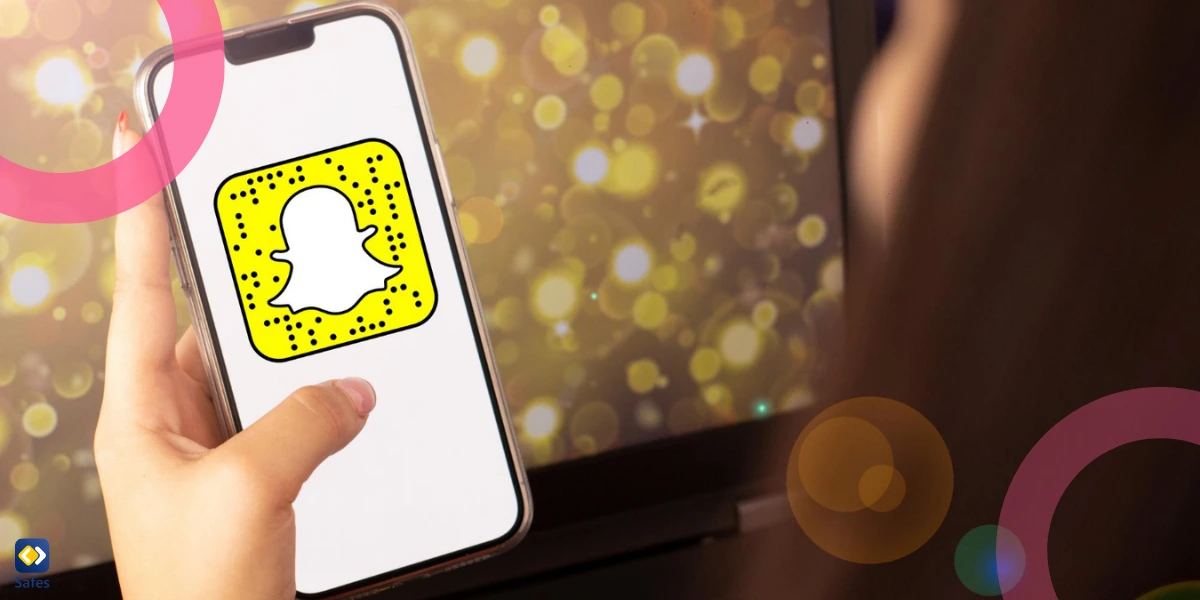 Eine Nahaufnahme einer Hand, die ein Smartphone hält, auf dessen Bildschirm das Snapchat-Logo angezeigt wird, mit einem Laptop und unscharfen goldenen Lichteffekten im Hintergrund.