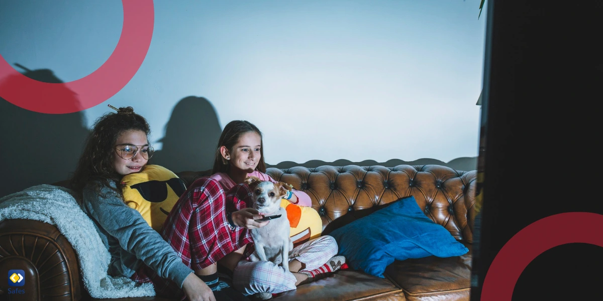 Two teenage girls in pajamas, with a dog, relax on a leather couch watching TV in a dimly lit room.