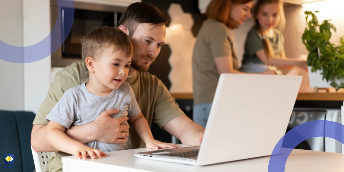 Vater und Sohn bedienen Laptop am Schreibtisch, während Mutter und Tochter im Hintergrund einer anderen Tätigkeit nachgehen.