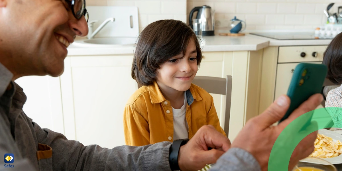 Ein Vater und sein Sohn sitzen zusammen an einem Esstisch in einer Küche. Der Vater, mit Brille und grauem Hemd, lächelt, als er seinem Sohn etwas auf einem grünen Smartphone zeigt