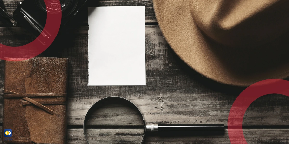 A flat lay of detective-themed items on a wooden table, including a camera, magnifying glass, leather notebook, felt hat, and a blank sheet of paper.