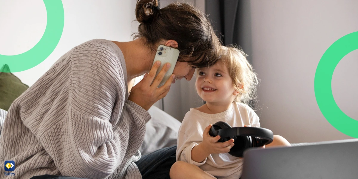 A mother playfully interacting with her toddler while holding a phone, as the child holds a pair of headphones near a laptop on a bed.