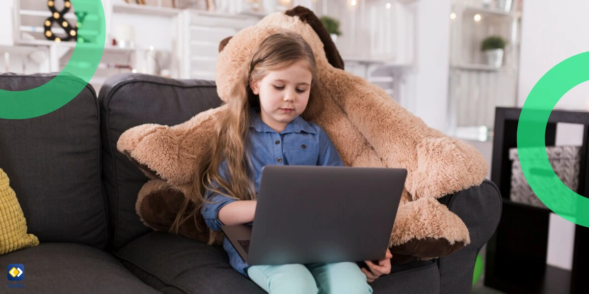 A young girl sitting on a couch, using a laptop while leaning against a large stuffed animal in a cozy living room setting.